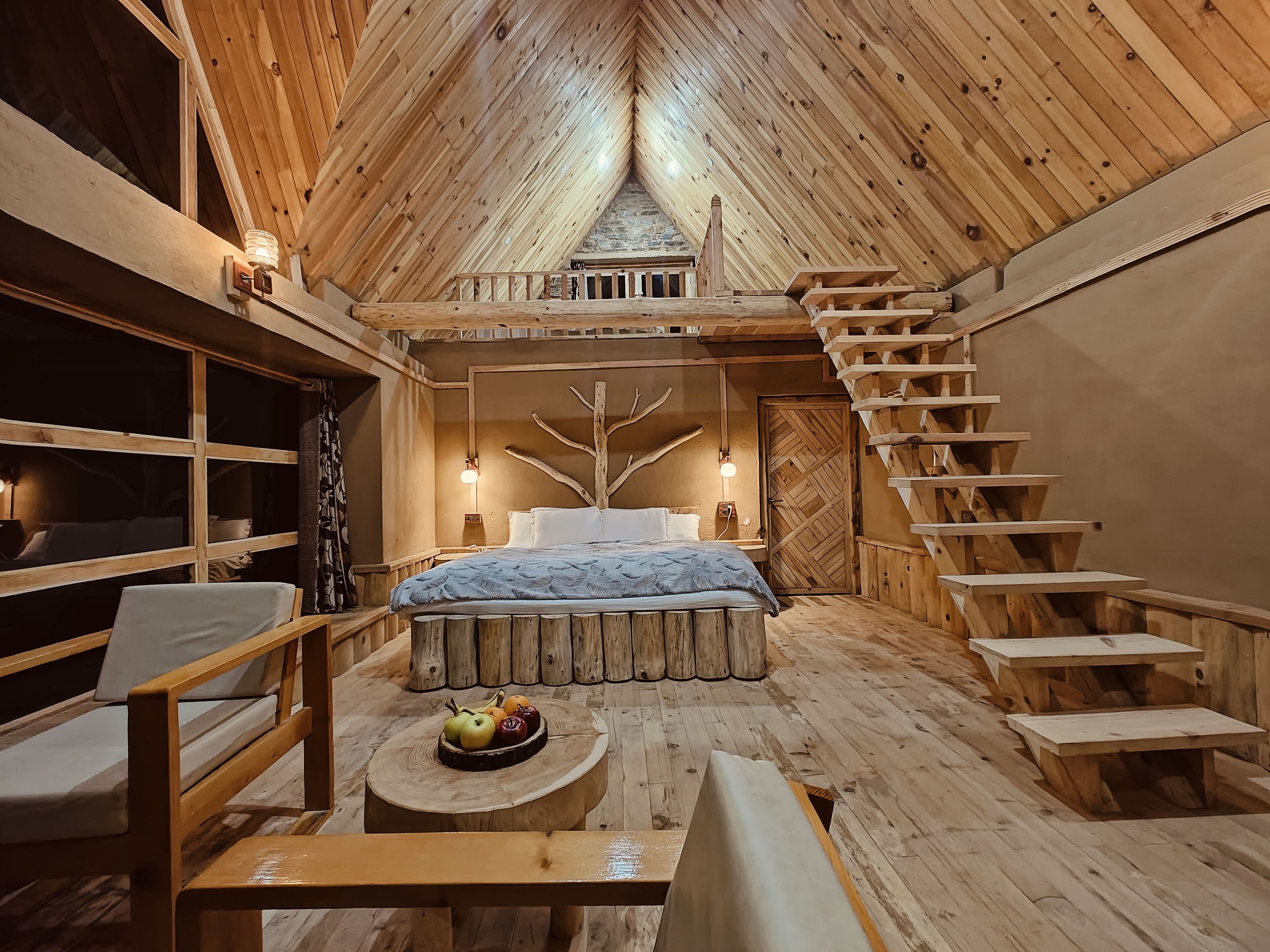 A cozy, wooden-themed bedroom with a bed, a small table with fruit, a bench, and a staircase.