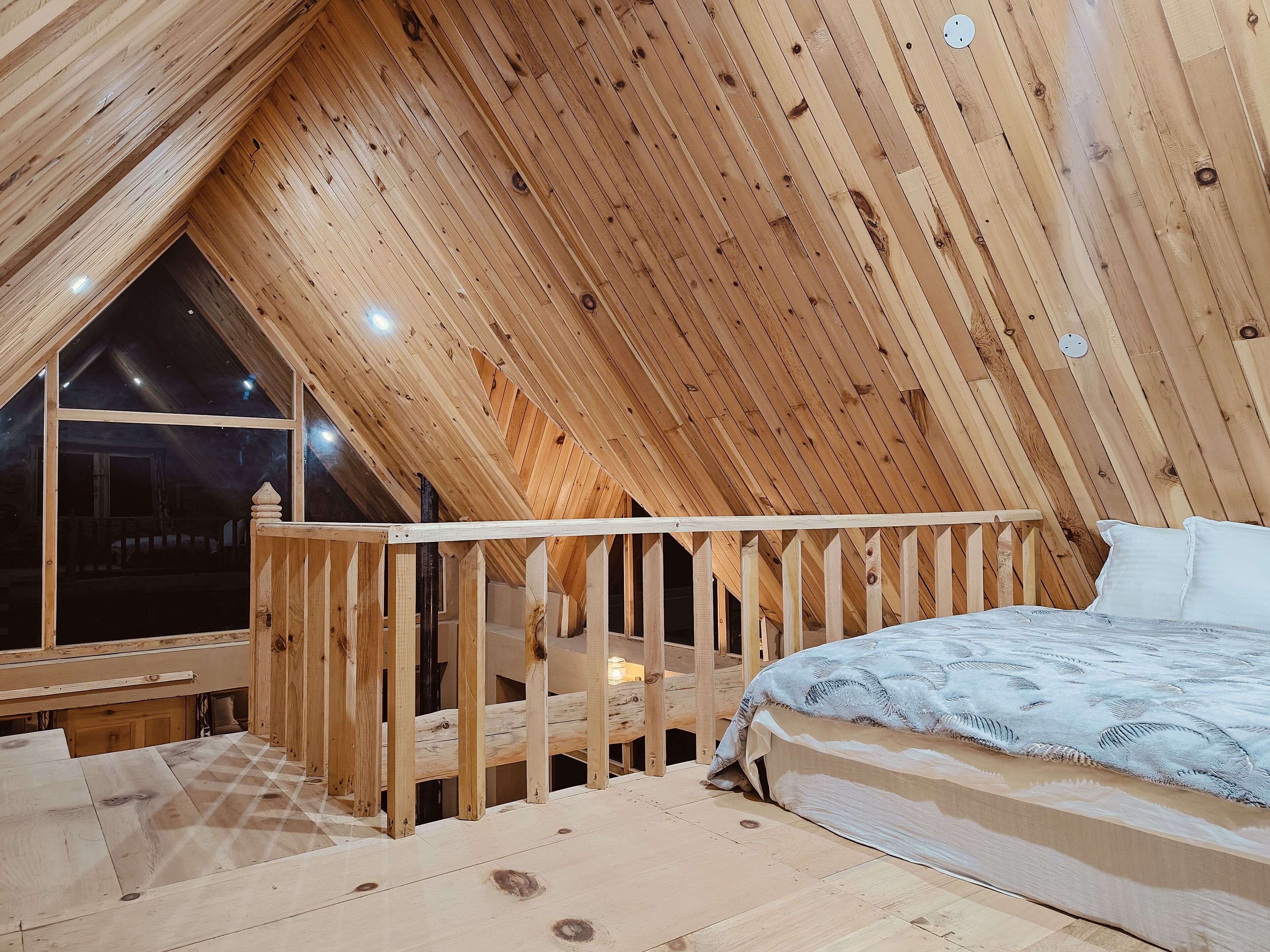 A bedroom with a sloped ceiling, wooden beams, a bed, and a window overlooking a dark exterior.