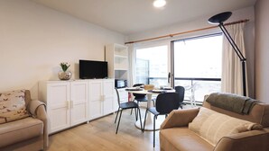 Una sala de estar moderna con un sofá, una mesa de comedor con sillas, un televisor y un ventanal con cortinas.