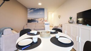 Un comedor con una mesa redonda dispuesta para cuatro personas, un sofá blanco y un televisor sobre un mueble.