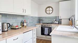 Une cuisine compacte avec des meubles blancs, une cuisinière à gaz, un four encastré, une bouilloire et une fenêtre ornée d’un vitrail décoratif.