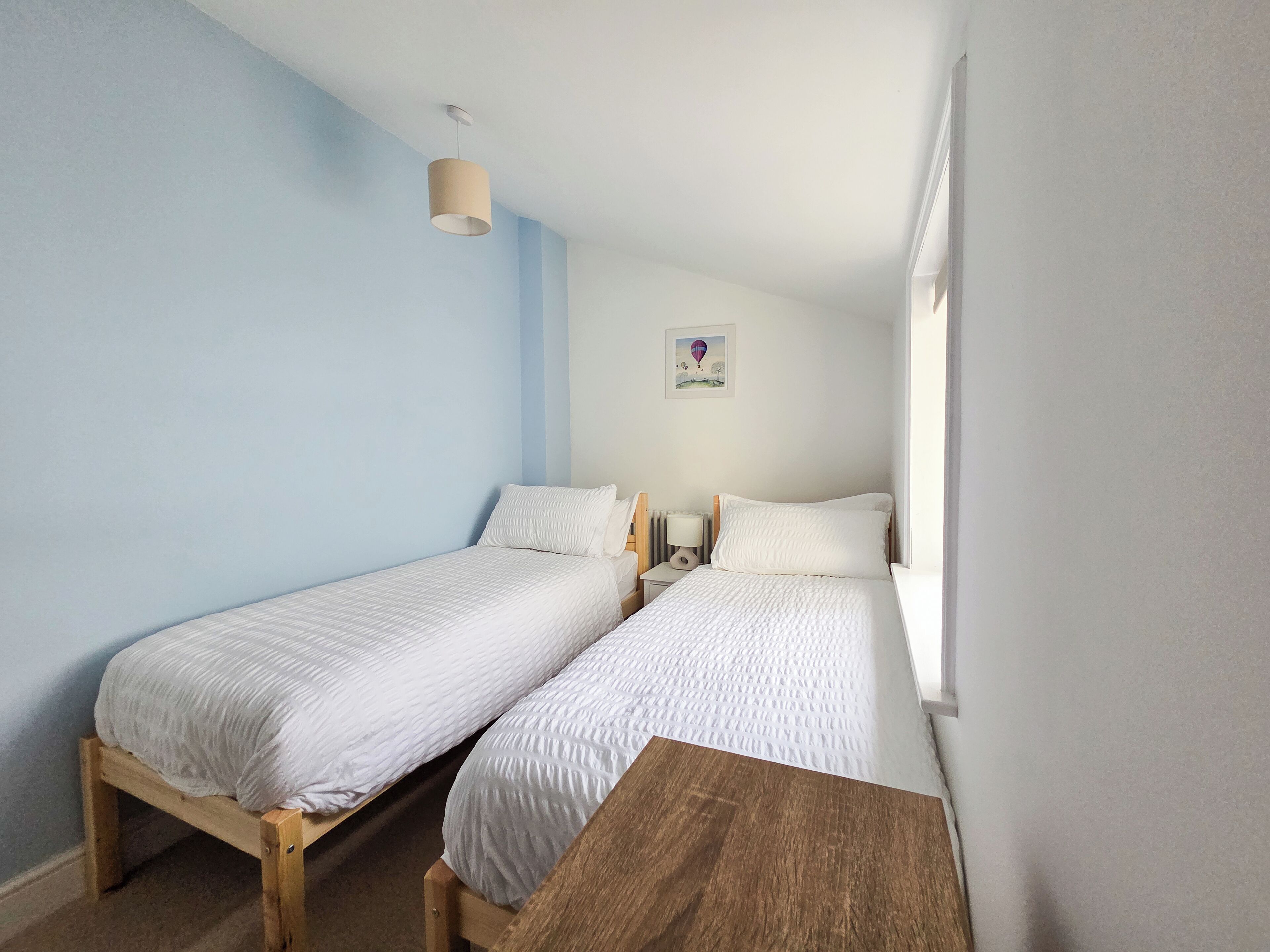 Une petite chambre propre, avec deux lits simples, une table de chevet en bois, une petite table et un luminaire suspendu.
