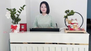 A reception desk with a woman seated behind it, a red clock, a decorative plant, and a cat figurine. - LOUIS OTIS APARTMENT (Da Nang)
