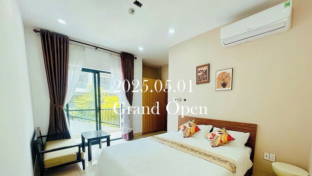 A hotel room with a large bed, a bedside table, a chair, a window with curtains, and an air conditioning unit on the wall. - LOUIS OTIS APARTMENT (Da Nang)