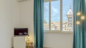A room with a TV, a small table with a lamp, teal curtains, and a window with a view of a building.
