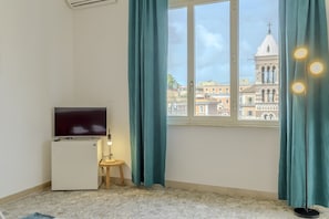 A room with a TV, a small table with a lamp, teal curtains, and a window with a view of a building. - Operetta View Apartment (ROMA)