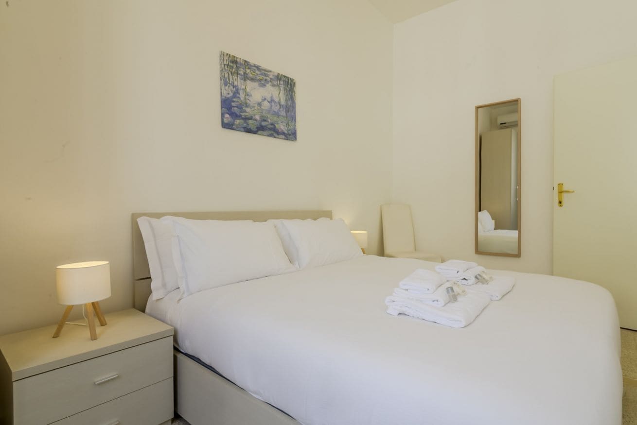 A hotel room with a bed, a nightstand with a lamp, a painting on the wall, and a mirror.
