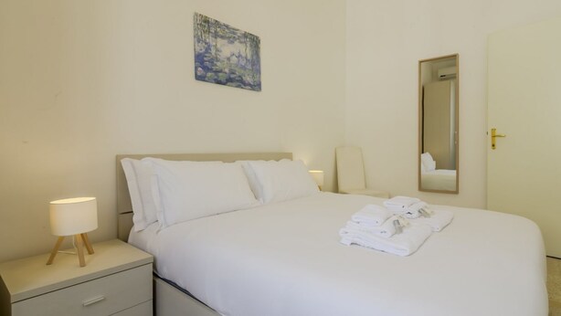A hotel room with a bed, a nightstand with a lamp, a painting on the wall, and a mirror.