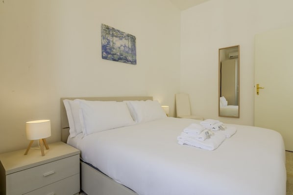 A hotel room with a bed, a nightstand with a lamp, a painting on the wall, and a mirror. - Operetta View Apartment (ROMA)