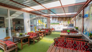 Une terrasse extérieure dotée de tables et de bancs en bois, recouverte d’un toit en métal et entourée de portes vitrées donnant accès à un bâtiment.