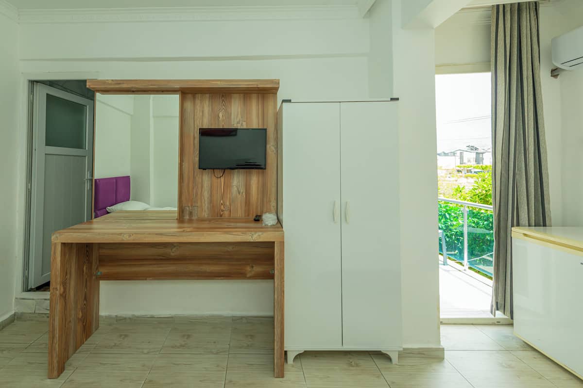 A room with a wooden desk, a white wardrobe, a television mounted on the wall, and a view of the outside balcony.