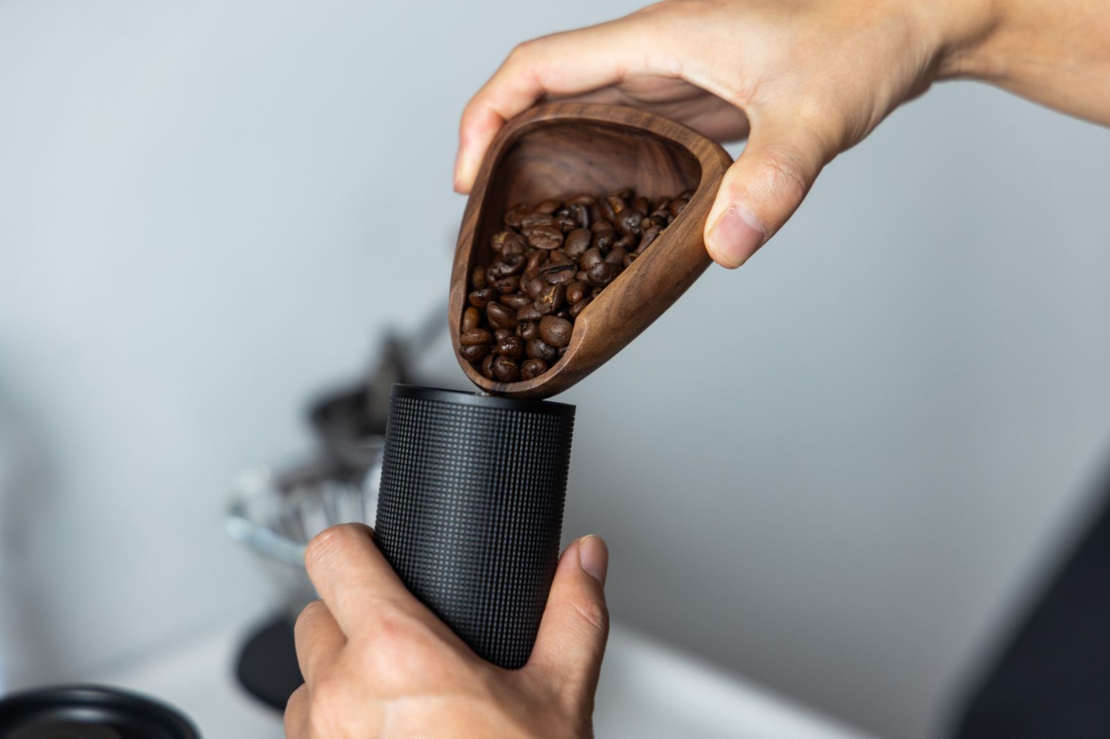 Una persona sostiene un cuenco de madera lleno de granos de café, vertiéndolos en un recipiente cilíndrico negro.