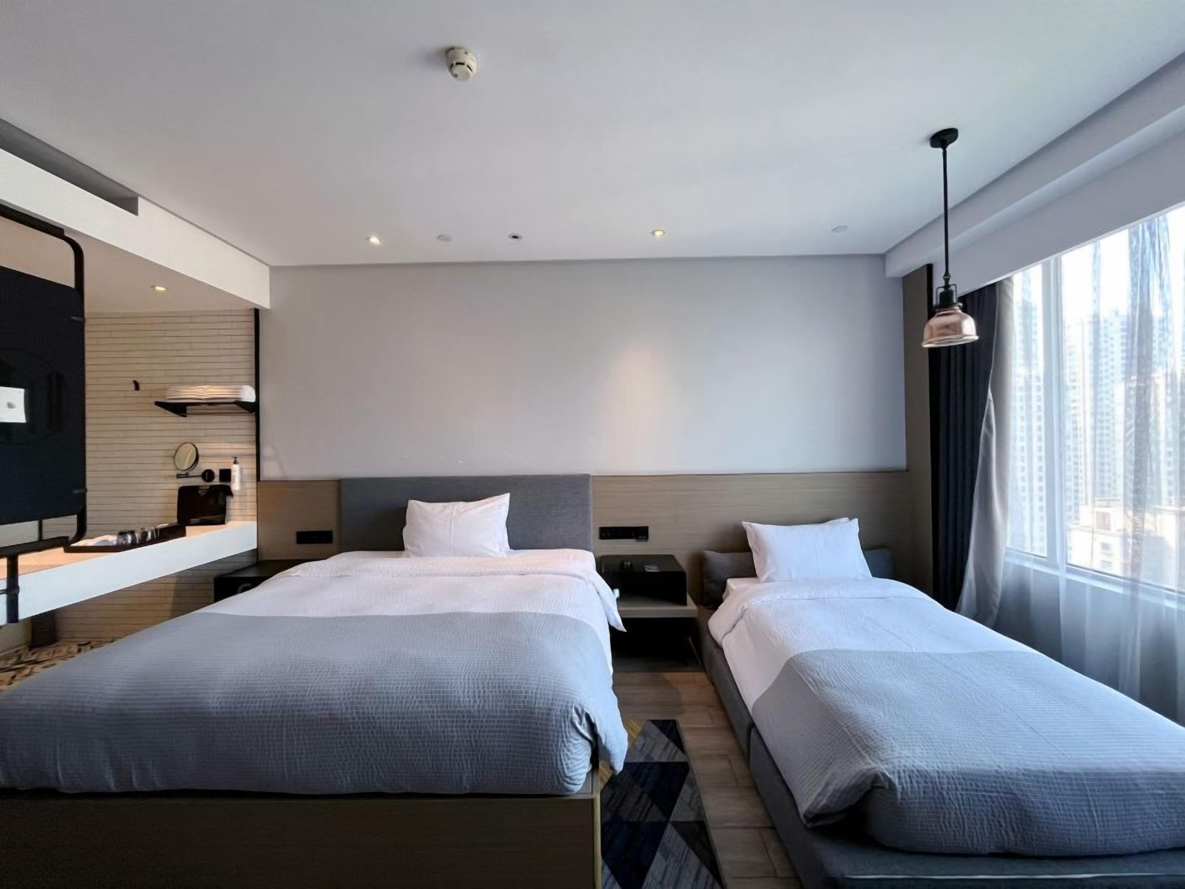 A modern hotel room with two beds, a large window with curtains, a flat-screen TV, and a small table with a lamp.