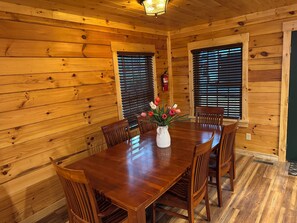 Un comedor de madera con una mesa, sillas, un jarrón de flores y ventanas con persianas.