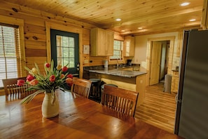 A wooden interior with a dining table, chairs, a vase of flowers, and a kitchen area with wooden cabinets and a refrigerator.