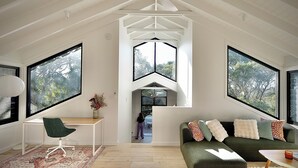 Un loft espacioso con techos altos, ventanas amplias que ofrecen vistas a árboles, un sofá verde y acogedor, un escritorio de madera y un tapete estampado.