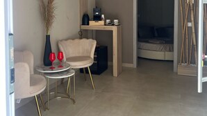 A modern living room with a sofa, a small round table with two red glasses, a tall vase with dried plants, a wooden desk with a computer and a coffee machine, and a bed with a dark headboard. - Charming deluxe suite 2 steps from the sea and downtown (Terrasini)