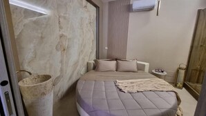 A modern bedroom with a marble wall, a round bed with a grey bedspread, a bedside table with a lamp, and a small nightstand. - Charming deluxe suite 2 steps from the sea and downtown (Terrasini)