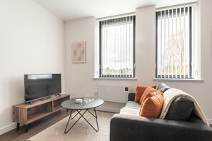 A modern living room with a flat-screen TV on a wooden stand, a round coffee table, and a grey sofa with orange cushions.