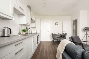 Una cocina moderna con armarios blancos, una zona de comedor con mesa y sillas, y un sofá gris.