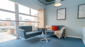 Un ufficio moderno con tappeto blu, un divano grigio con cuscini rossi e beige, un tavolino e ampie finestre che offrono vista sugli edifici esterni.