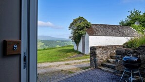 A view from a room with a door leading to a gravel path, a stone wall, and a white building with a tiled roof. - Cottage in Conwy with Hot Tub & Mountain Views (Eglwysbach)