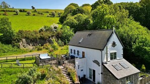 A white building with a stone foundation, a sloped roof, and a small deck with outdoor seating. - Cottage in Conwy with Hot Tub & Mountain Views (Eglwysbach)