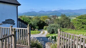 A rural landscape with a white building, a wooden fence, and a gravel path leading to a green valley. - Cottage in Conwy with Hot Tub & Mountain Views (Eglwysbach)
