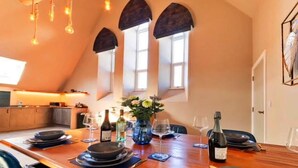 A dining area with a long wooden table set for six, flanked by arched windows and a kitchen in the background. - Chapel in Newquay near Towan Beach (Newquay)