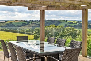 A glass-top table with wicker chairs in a covered outdoor area with a scenic view of rolling hills and fields. - Cottage in Whitestone with Garden & Games Room (Whitestone)