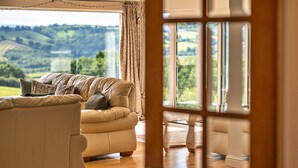 A cozy living room with a beige sofa, a wooden coffee table, and a view of rolling hills through large windows.