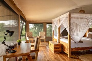 A room with a wooden desk, two chairs, a bedside table, a bed with a canopy, and a window with a view of trees. - Manya Luxury Camp (Serengeti)