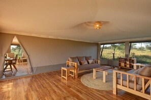 A spacious room with a large window, a sofa, a coffee table, and a wooden floor. - Manya Luxury Camp (Serengeti)