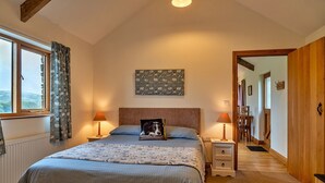 A bedroom with a bed, two bedside tables with lamps, a window with curtains, a wooden door, and a wall-mounted artwork. - Countryside cottage with private hot tub, No Deposit (Liskeard)