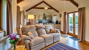 A cozy living room with a sofa, a coffee table with a vase of flowers, and a view of the kitchen and outdoor area through a glass door. - Countryside cottage with private hot tub, No Deposit (Liskeard)