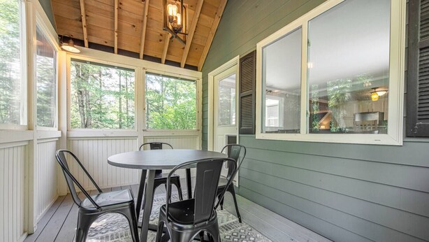 A covered porch with a round table and four chairs, wooden ceiling beams, and a view of a kitchen through a window. - Beautiful White Mountains Home w/ Sauna, Firepit and  Gameroom Local Skiing, Hiking and Golf. (Thornton)