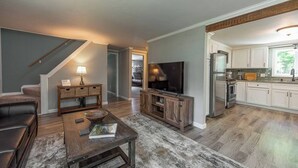 A modern living room with a sofa, coffee table, and TV stand. A staircase leads to another level. The kitchen features white cabinets, a refrigerator, and a window. - Beautiful White Mountains Home w/ Sauna, Firepit and  Gameroom Local Skiing, Hiking and Golf. (Thornton)
