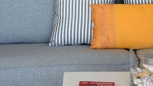 A living room with a grey sofa, striped pillows, a yellow cushion, a glass vase with seashells, and a lamp on a wooden side table.