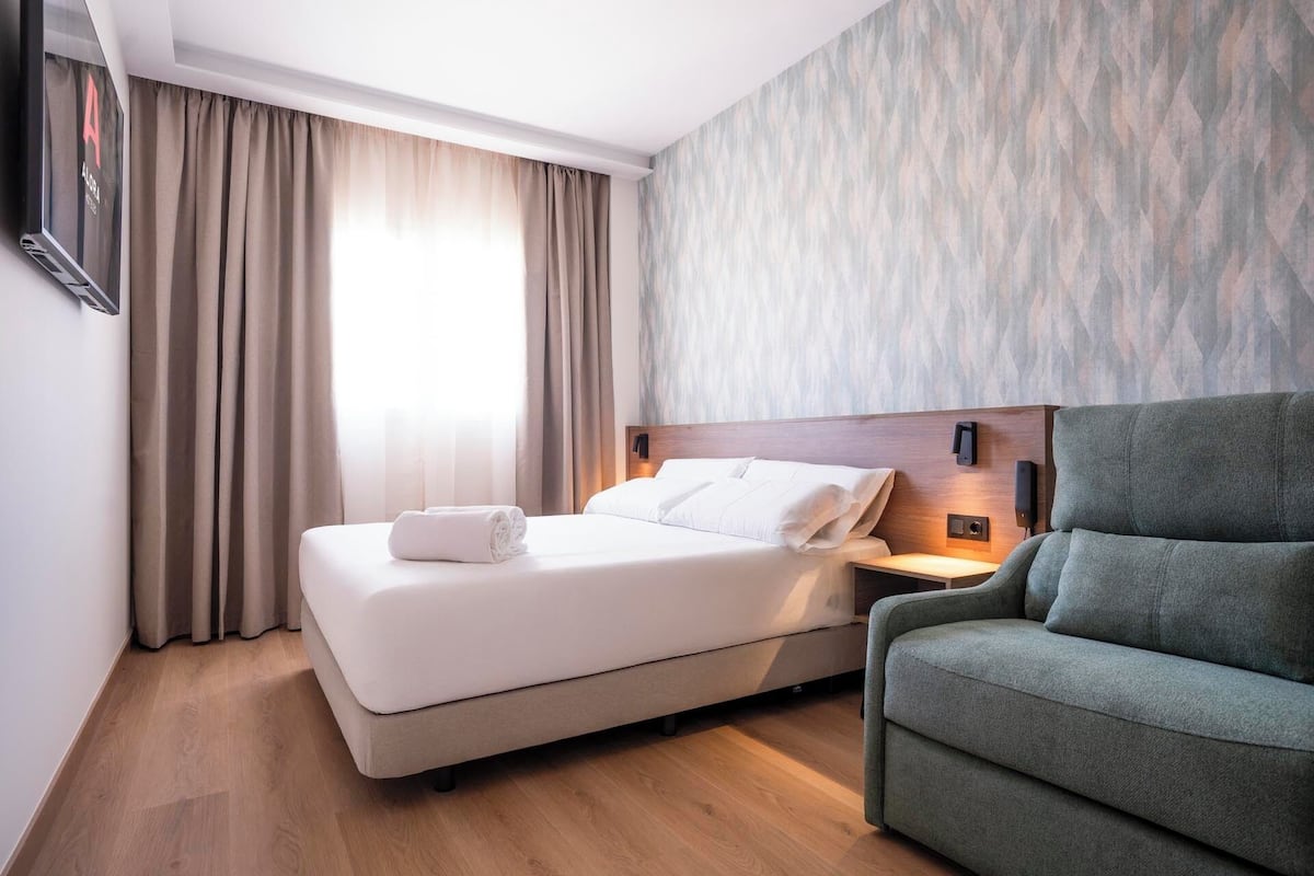 A hotel room with a bed, a green armchair, a nightstand with a lamp, and a wall-mounted TV.