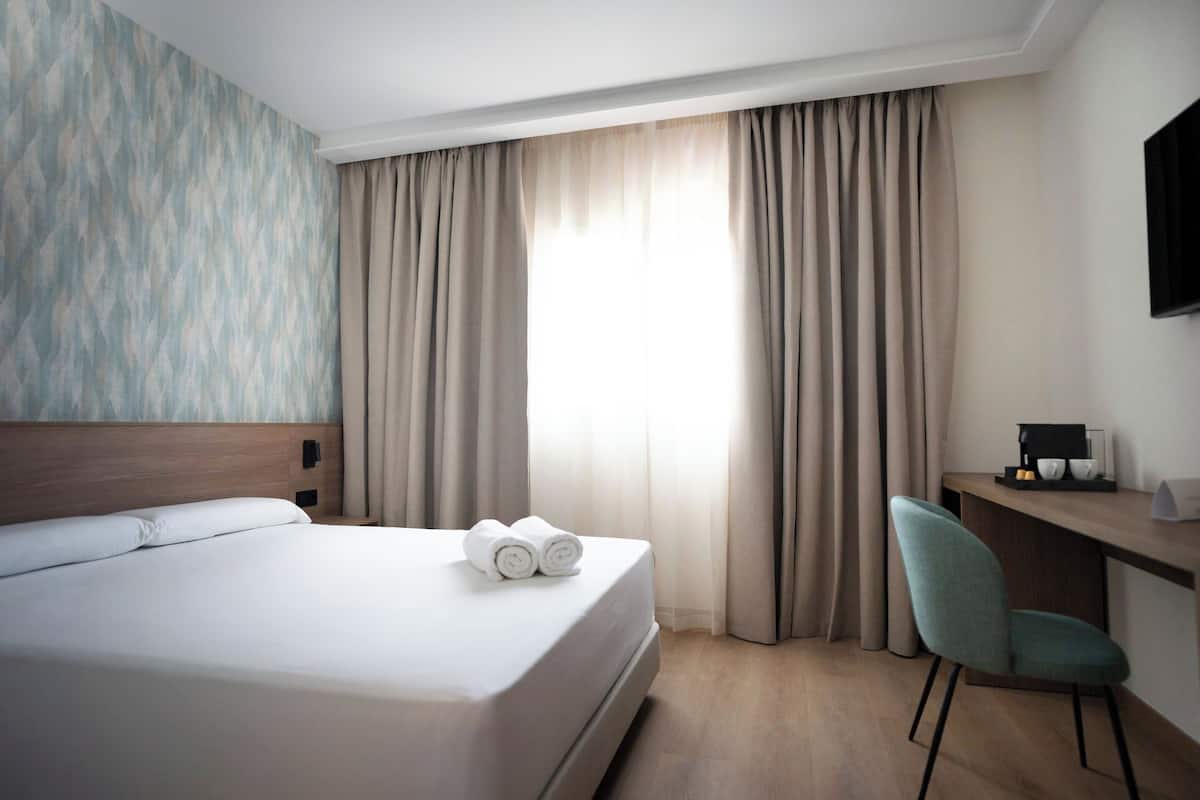 A hotel room with a bed, a desk, a chair, and a window with curtains.
