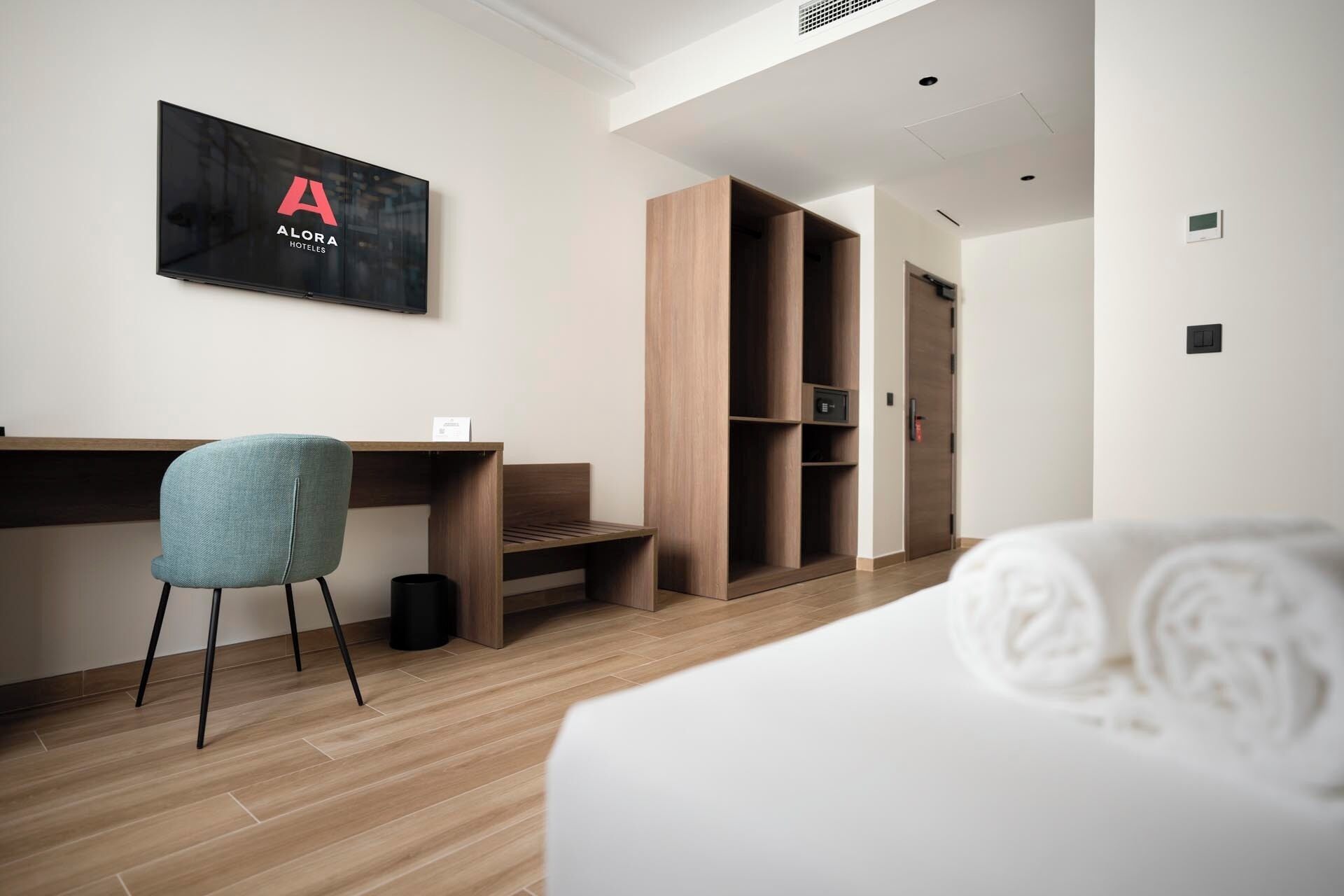 Une chambre d’hôtel équipée d’un lit, d’un bureau, d’une chaise, d’une armoire et d’un téléviseur fixé au mur, sur lequel est affiché le nom de l’hôtel.