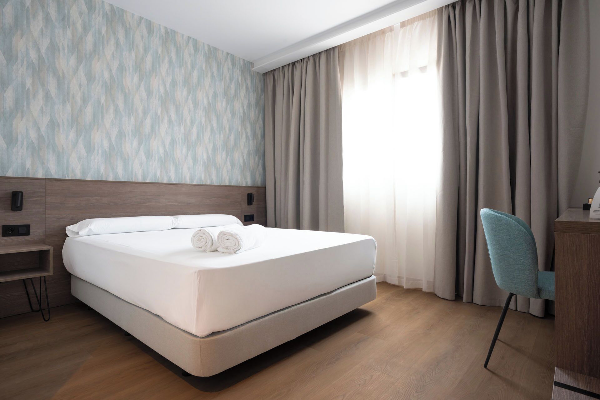 A hotel room with a bed, a desk, a chair, and a window with curtains.