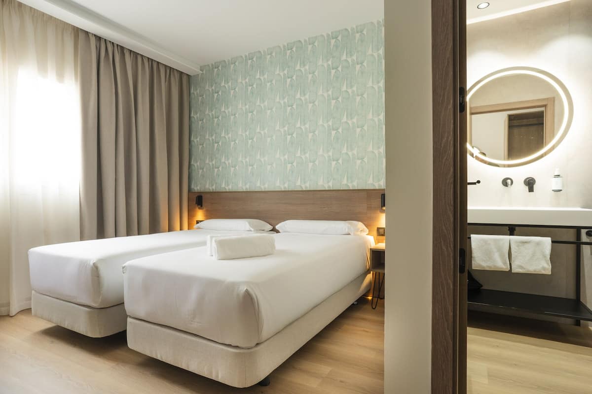 A hotel room with two beds, a bathroom with a mirror and sink, and a light-colored wooden floor.