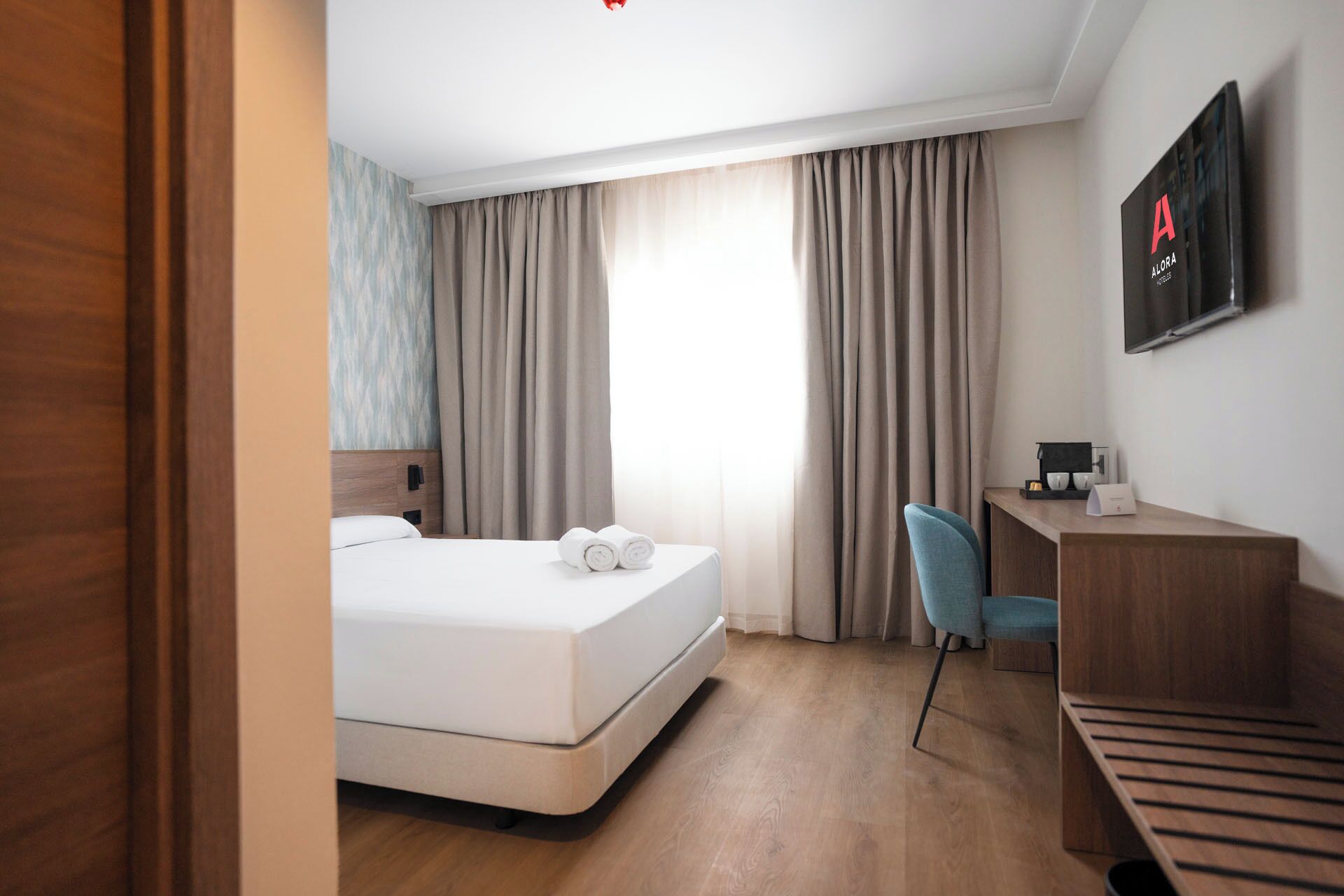 A hotel room with a bed, a desk, a chair, and a television on the wall.