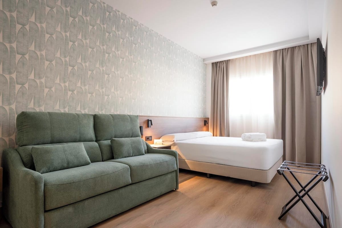A hotel room with a green sofa, a bed with white linens, a nightstand with a lamp, and a window with curtains.