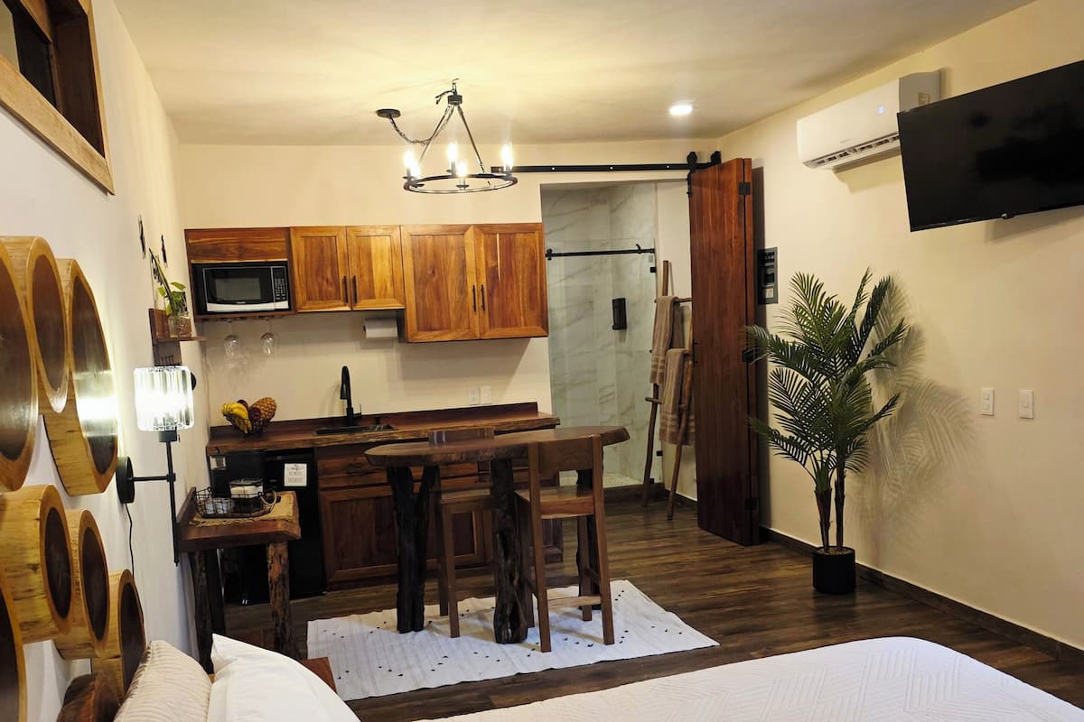 A bedroom with a large bed, a wooden dining table, and a kitchen area with wooden cabinets and a microwave.