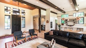 A spacious living area with a black leather sofa, a coffee table, and a dining table with chairs. There is a kitchen in the background with white cabinets and a large window. - This idyllic Suncadia home features a speakeasy, cowboy cold dip, outdoor shower, private library, chef's kitchen, golf course views, and MORE (Cle Elum)