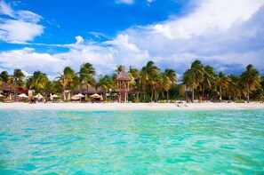 A beach with clear turquoise water, palm trees, and a thatched-roof structure.