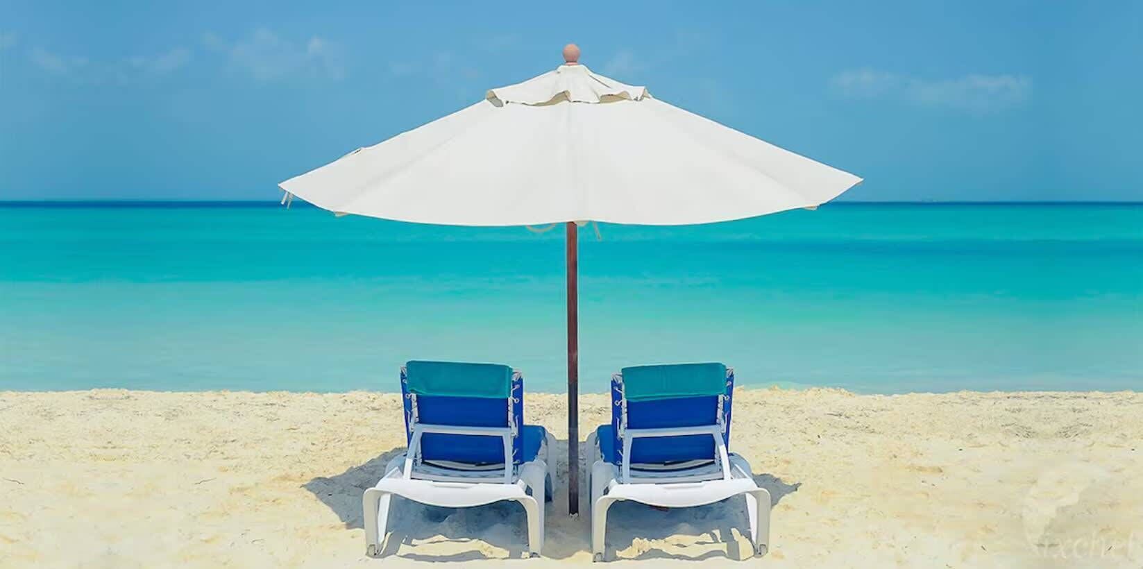 Two beach chairs with a white umbrella on a sandy beach with clear turquoise water.
