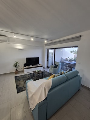 A modern living room with a sofa, TV, and a dining area visible through a glass door.